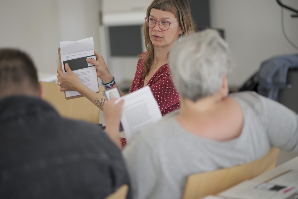 Inga hält das Klemmbrett und zeigt die Nutzung der Lesehilfe. Im Vordergrund sind verschwommene Teilnehmende.
